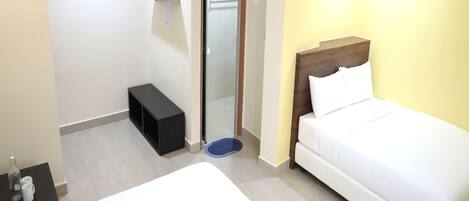 A hotel room with a bed, a small bedside table with a bottle and two cups, a black storage unit, a mirror, and a door leading to a bathroom.
