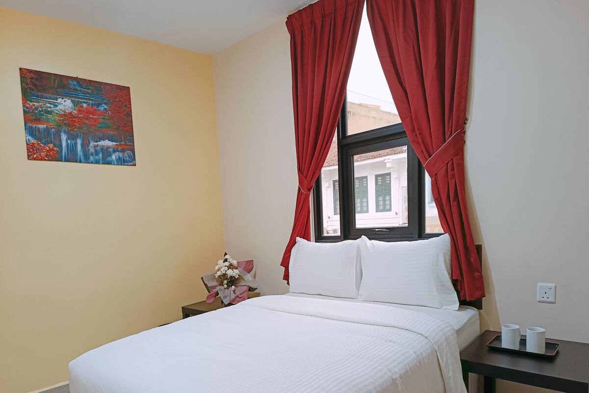 A hotel room with a bed, a nightstand, a window with red curtains, a painting on the wall, and a view of buildings outside.