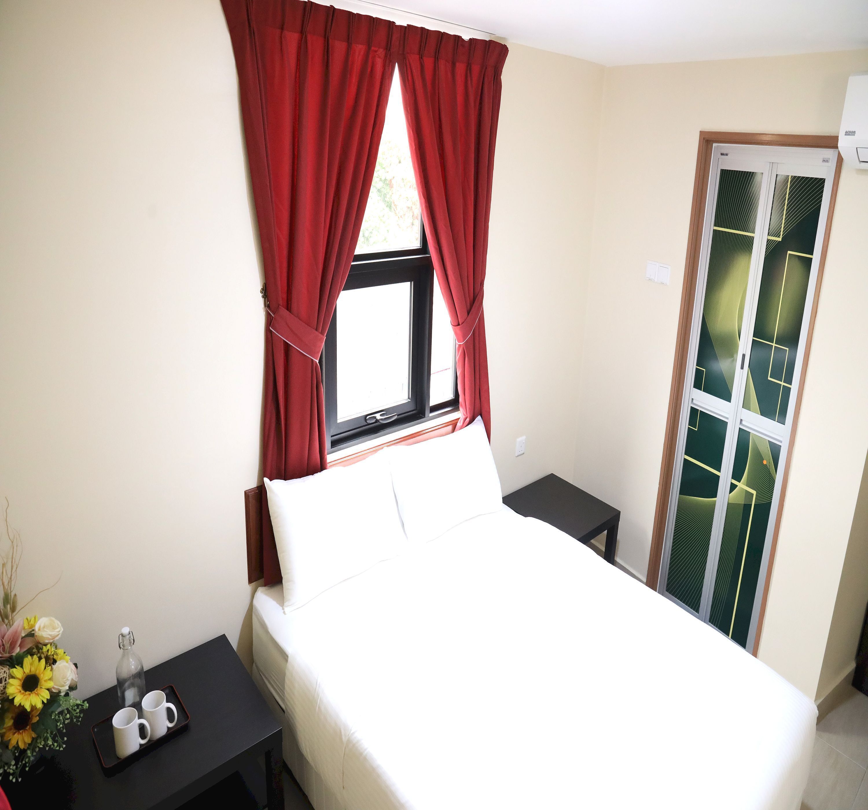 A hotel room with a bed, a nightstand, a window with red curtains, a door with a geometric glass design, and a small table with two white mugs.