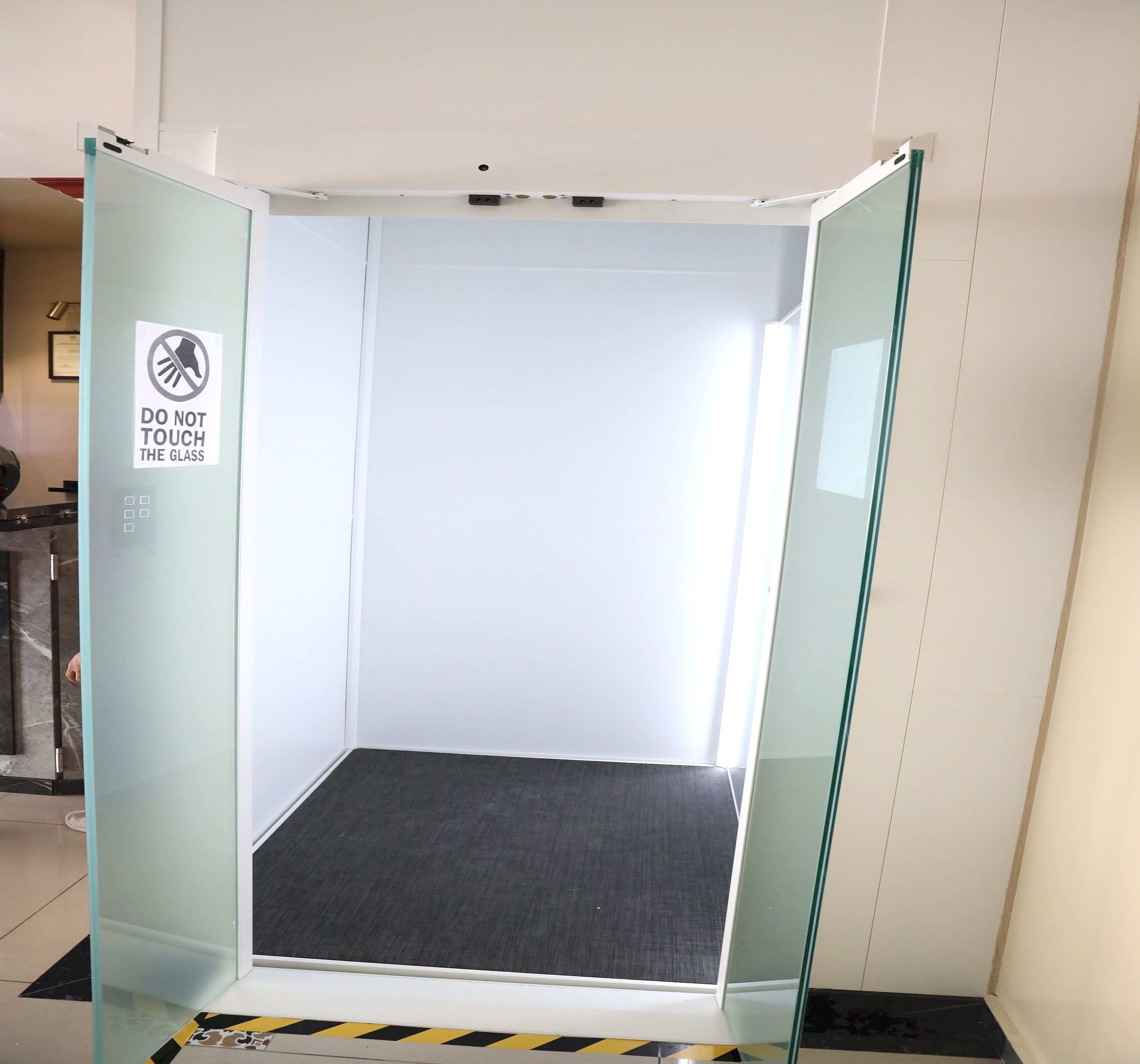 A glass-walled elevator with a "DO NOT TOUCH THE GLASS" sign, a control panel with buttons, and a black mat at the bottom.