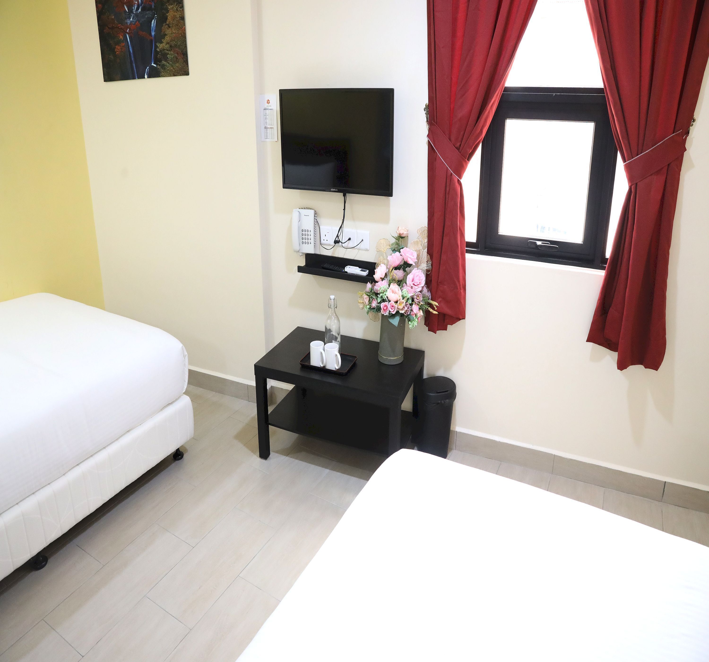 A hotel room with a bed, a small table with a vase of flowers, a TV, a phone, and a window with red curtains.