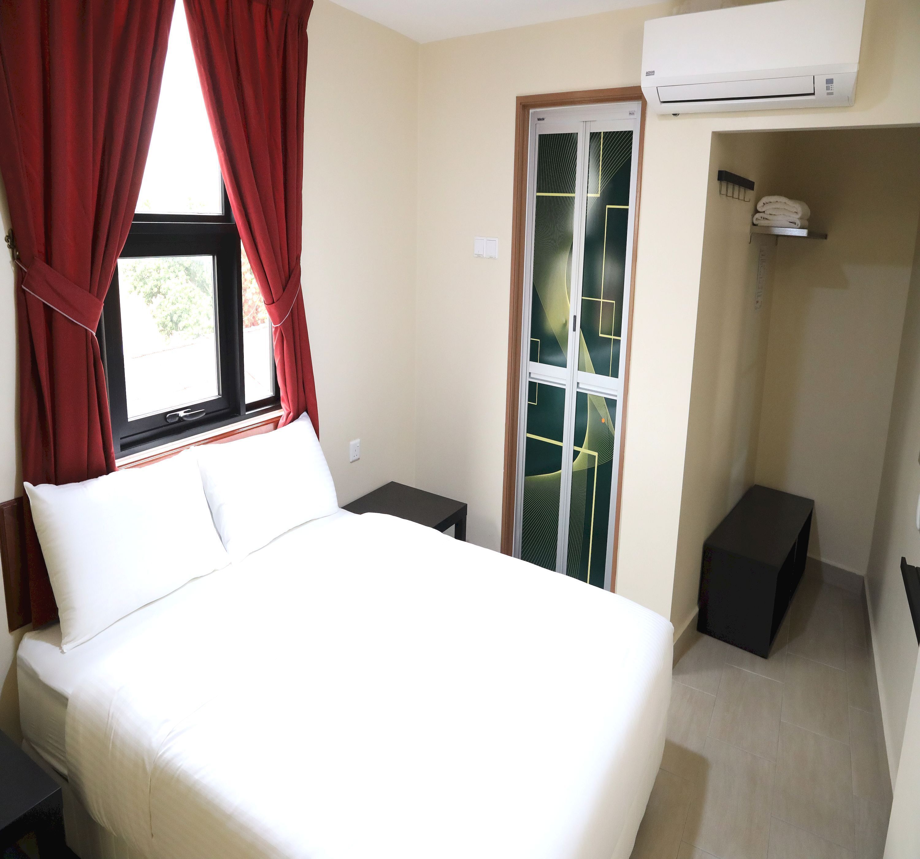 A hotel room with a bed, a window with red curtains, a door with a geometric pattern, an air conditioning unit, and a small black table.