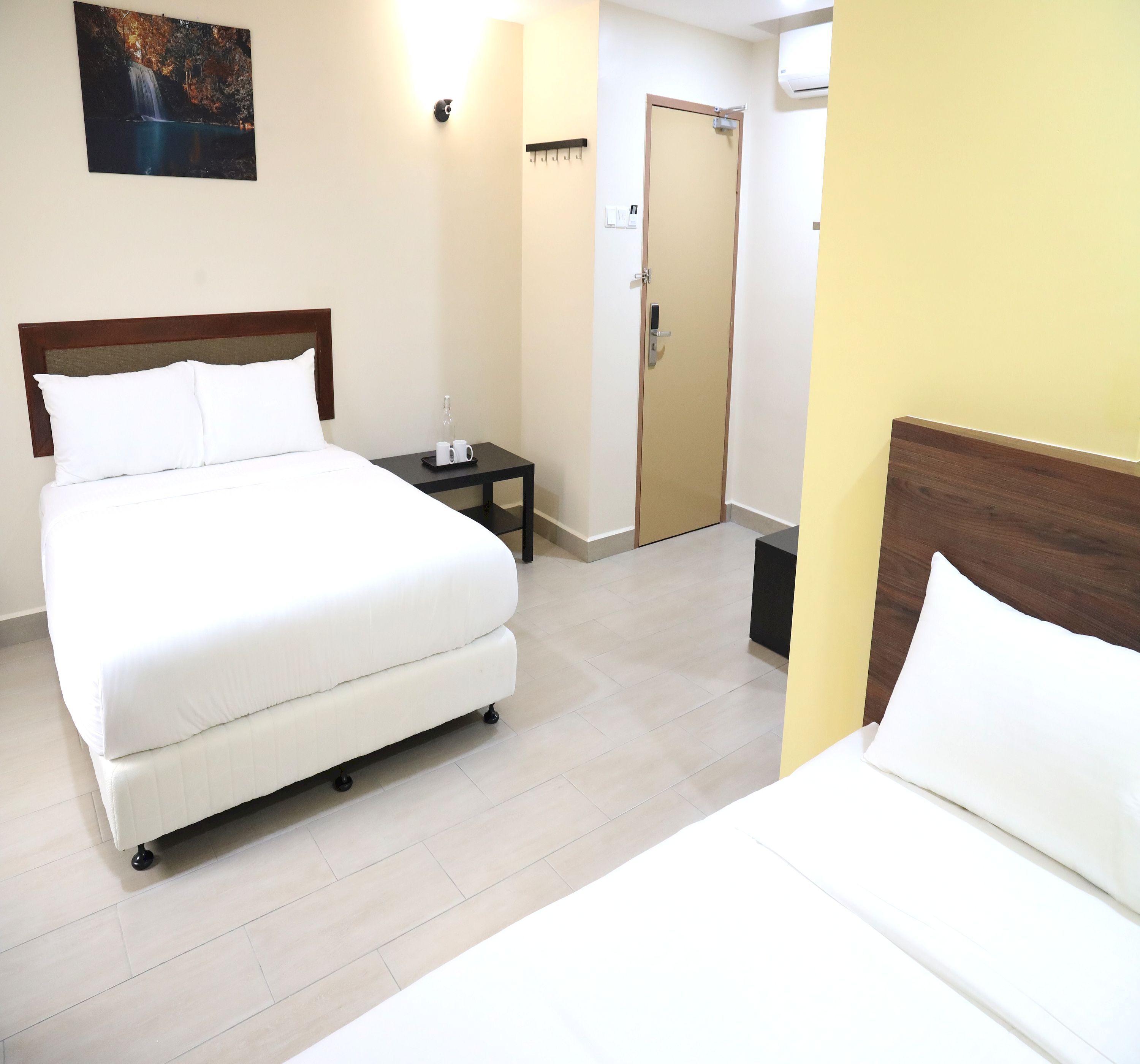 A hotel room with a large bed, a bedside table with a bottle and glasses, a door, and a wall-mounted light.