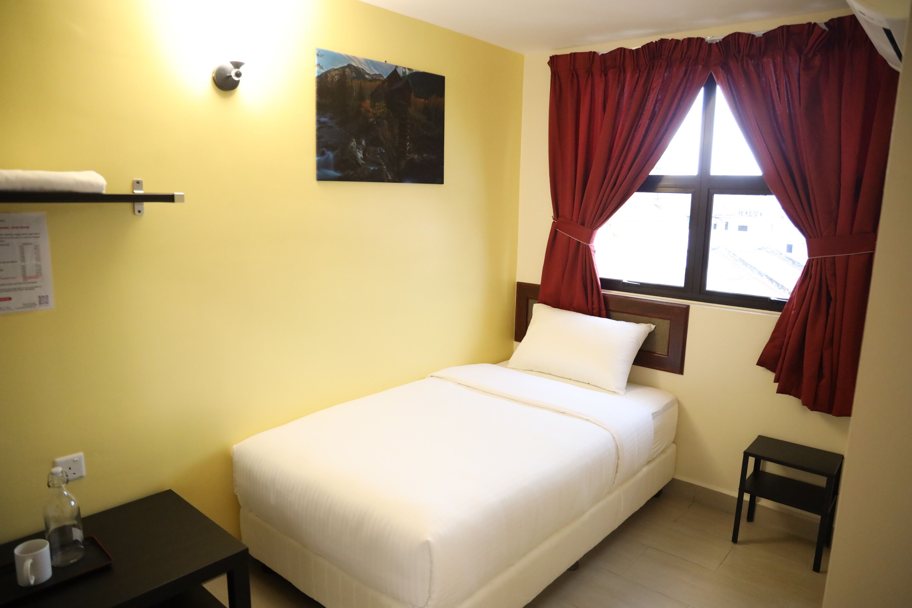 Una cama individual con ropa de cama blanca en una habitación con paredes amarillas, un cuadro y una ventana con cortinas rojas.