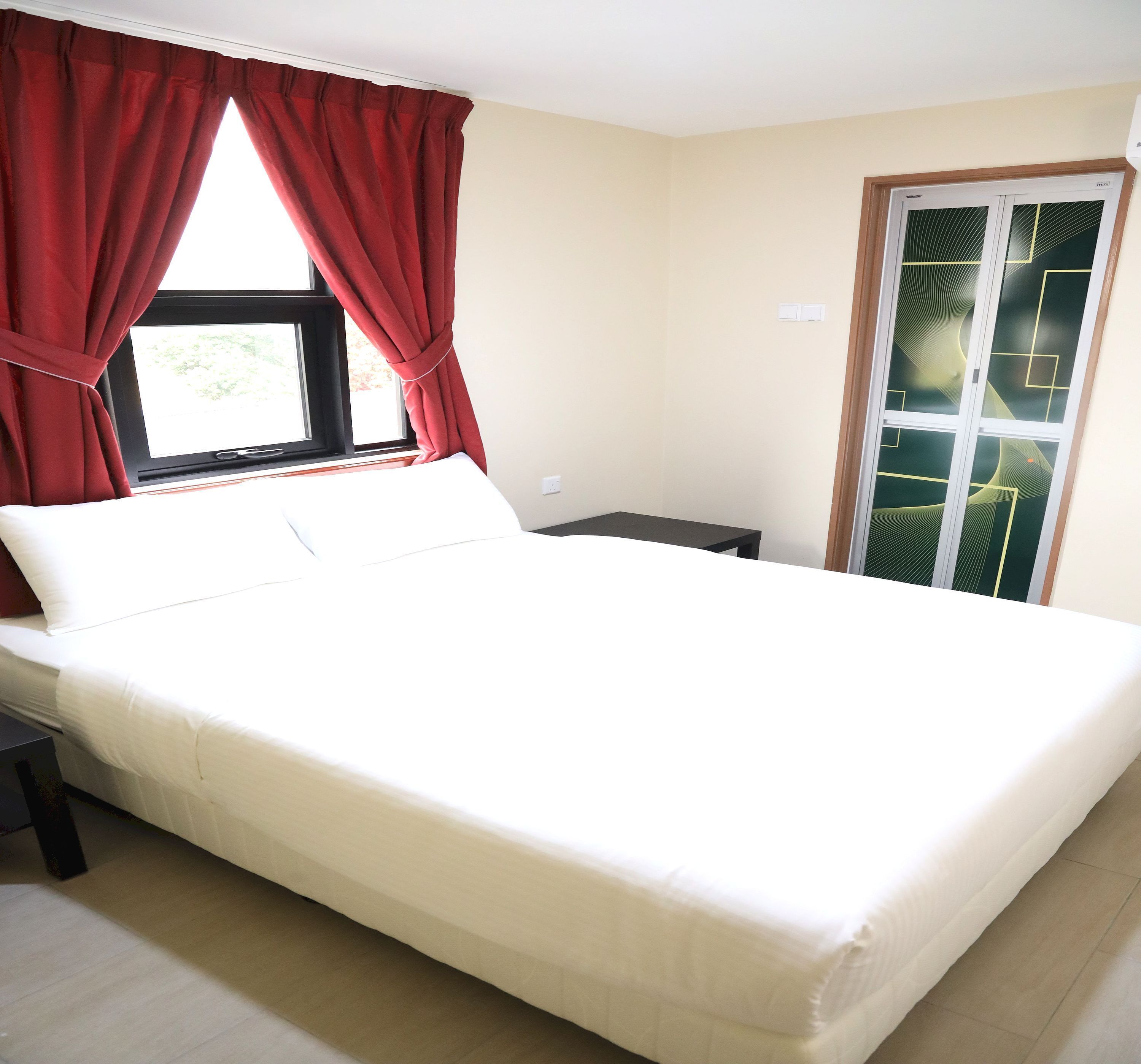 Un dormitorio con cama, una ventana con cortinas rojas, una mesita de noche y una puerta con diseños geométricos.
