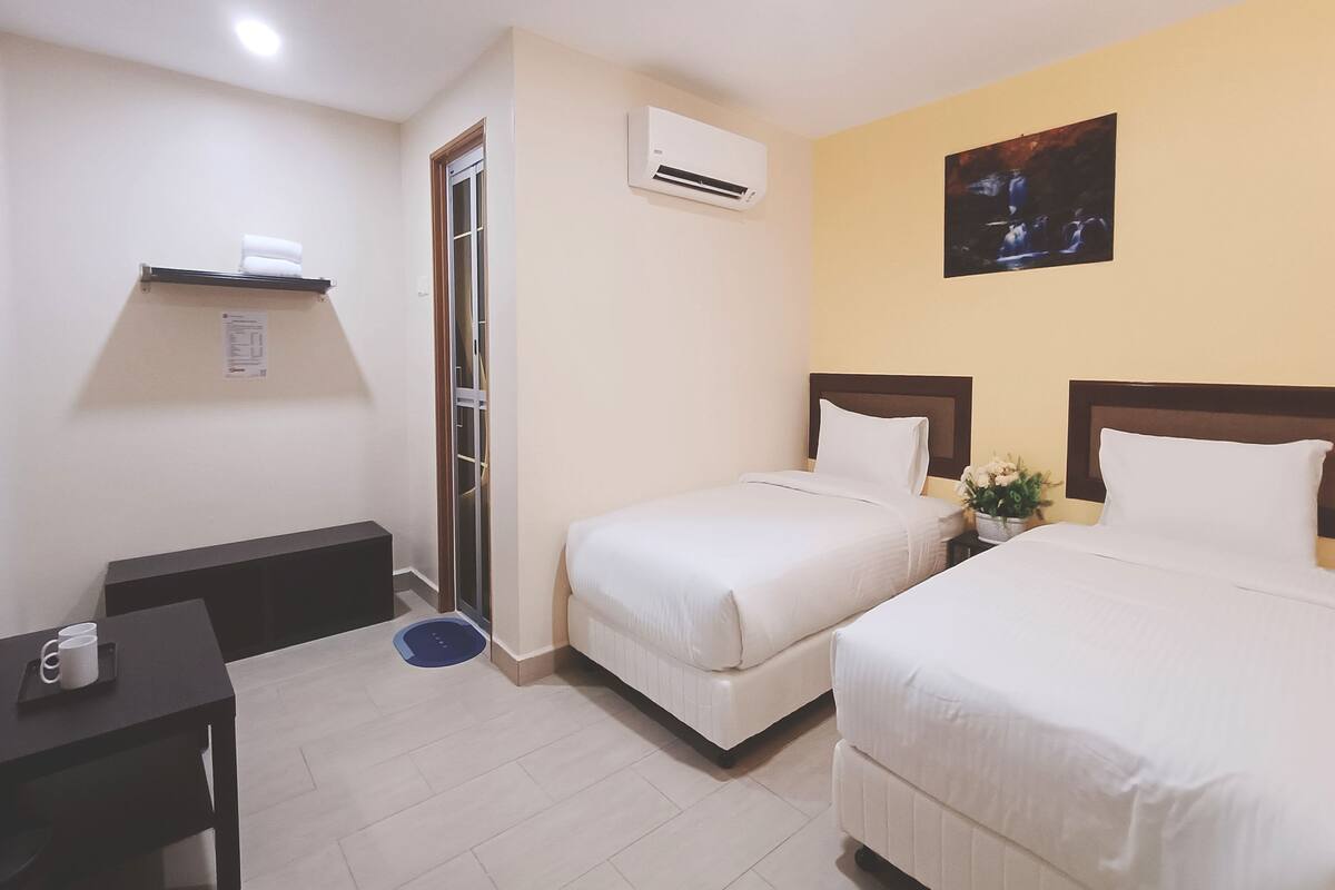 A hotel room with two beds, a desk, a small table, a trash bin, an air conditioning unit, and a painting on the wall.