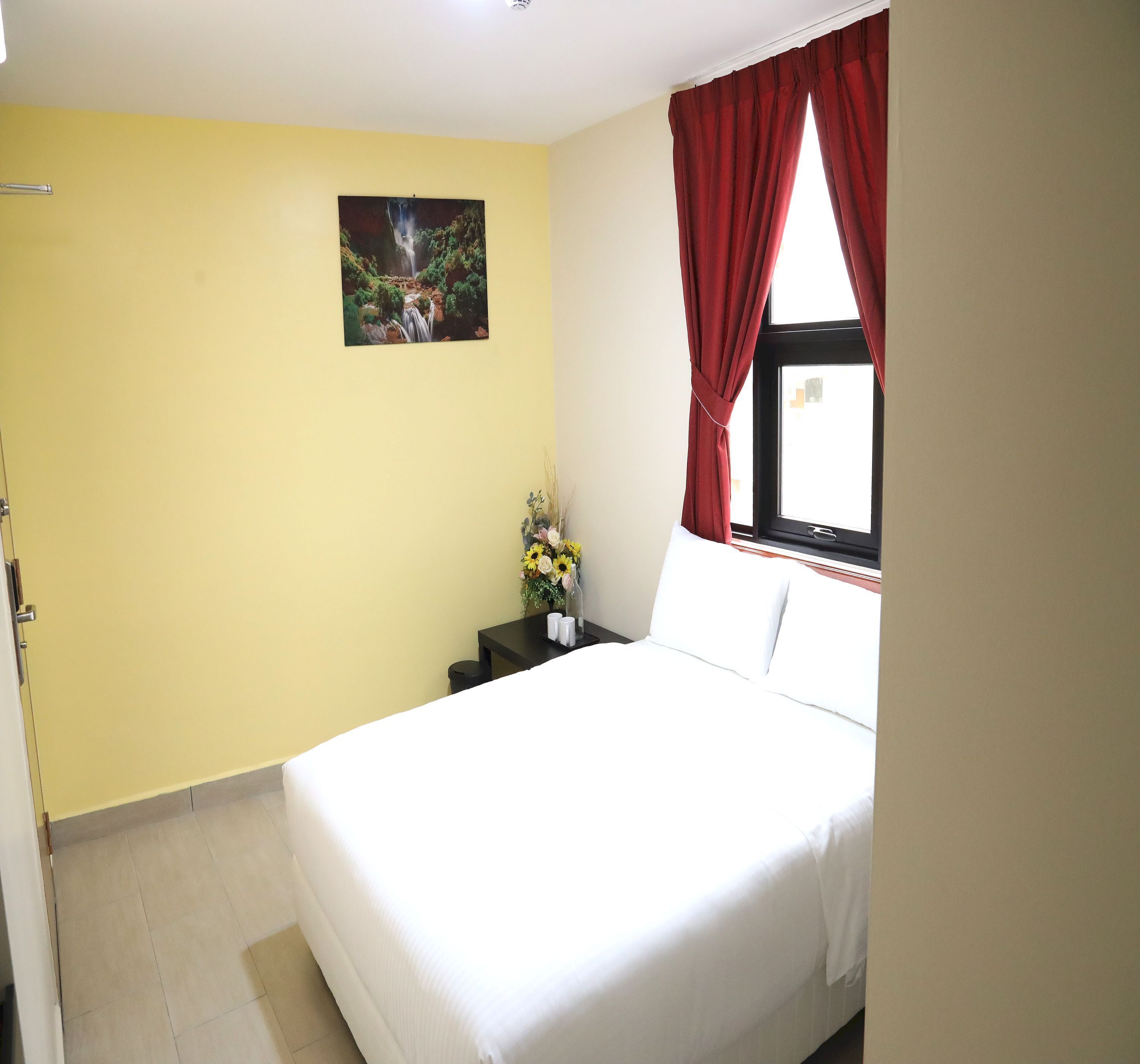 A hotel room with a bed, a nightstand with a vase of flowers, a painting on the wall, and a window with red curtains.