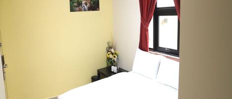 A hotel room with a bed, a nightstand with a vase of flowers, a painting on the wall, and a window with red curtains.