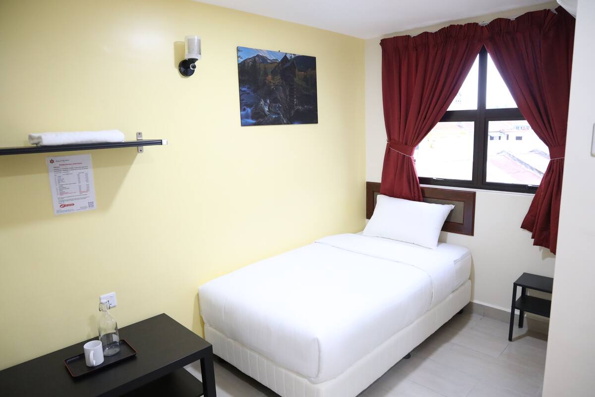 A neatly arranged hotel room with a bed, a small table with a tray, a wall-mounted shelf, a window with red curtains, and a painting on the wall.