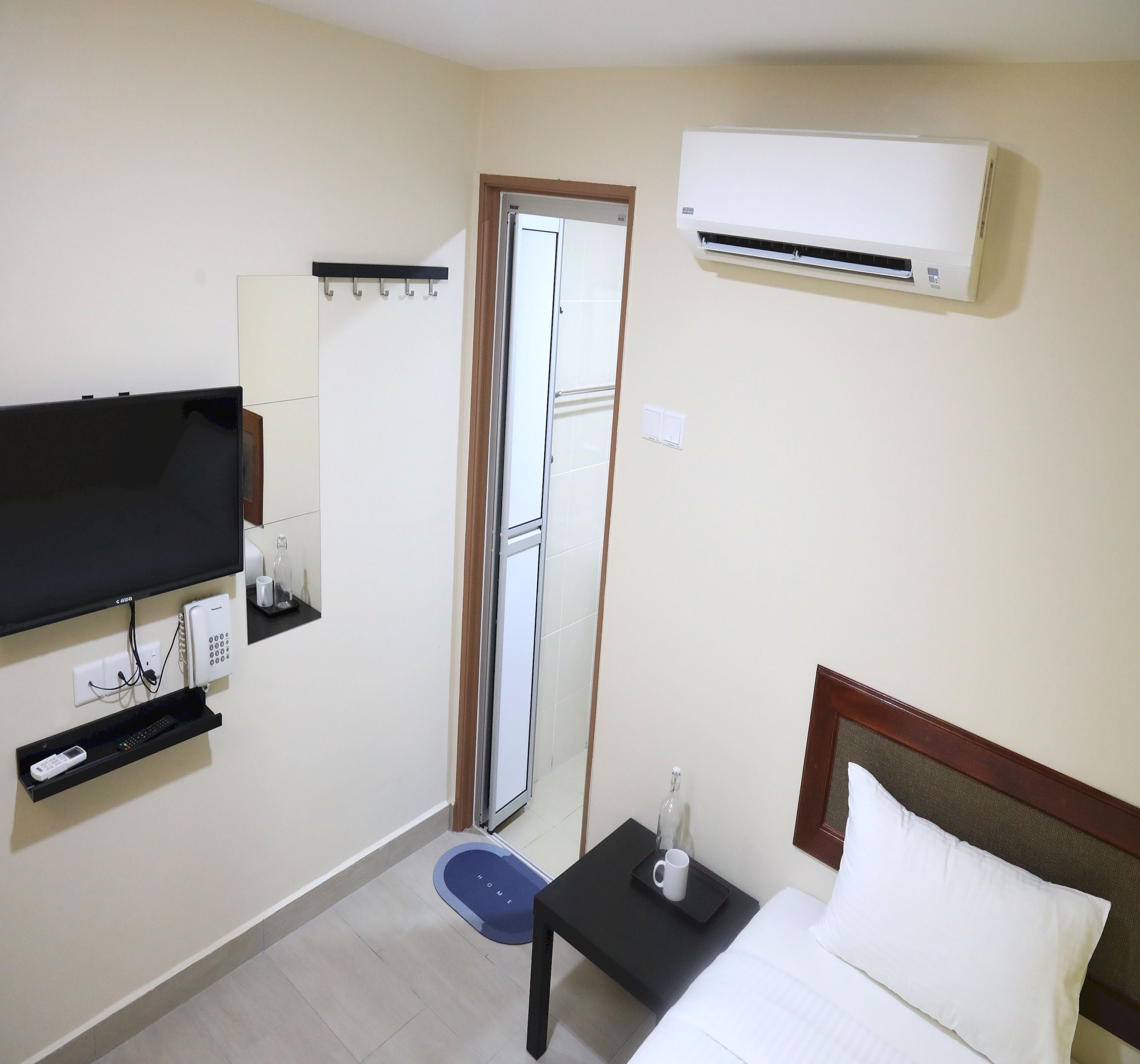 A hotel room with a bed, a TV mounted on the wall, a small table with a remote control, a wall-mounted air conditioner, and a door with a mirror.