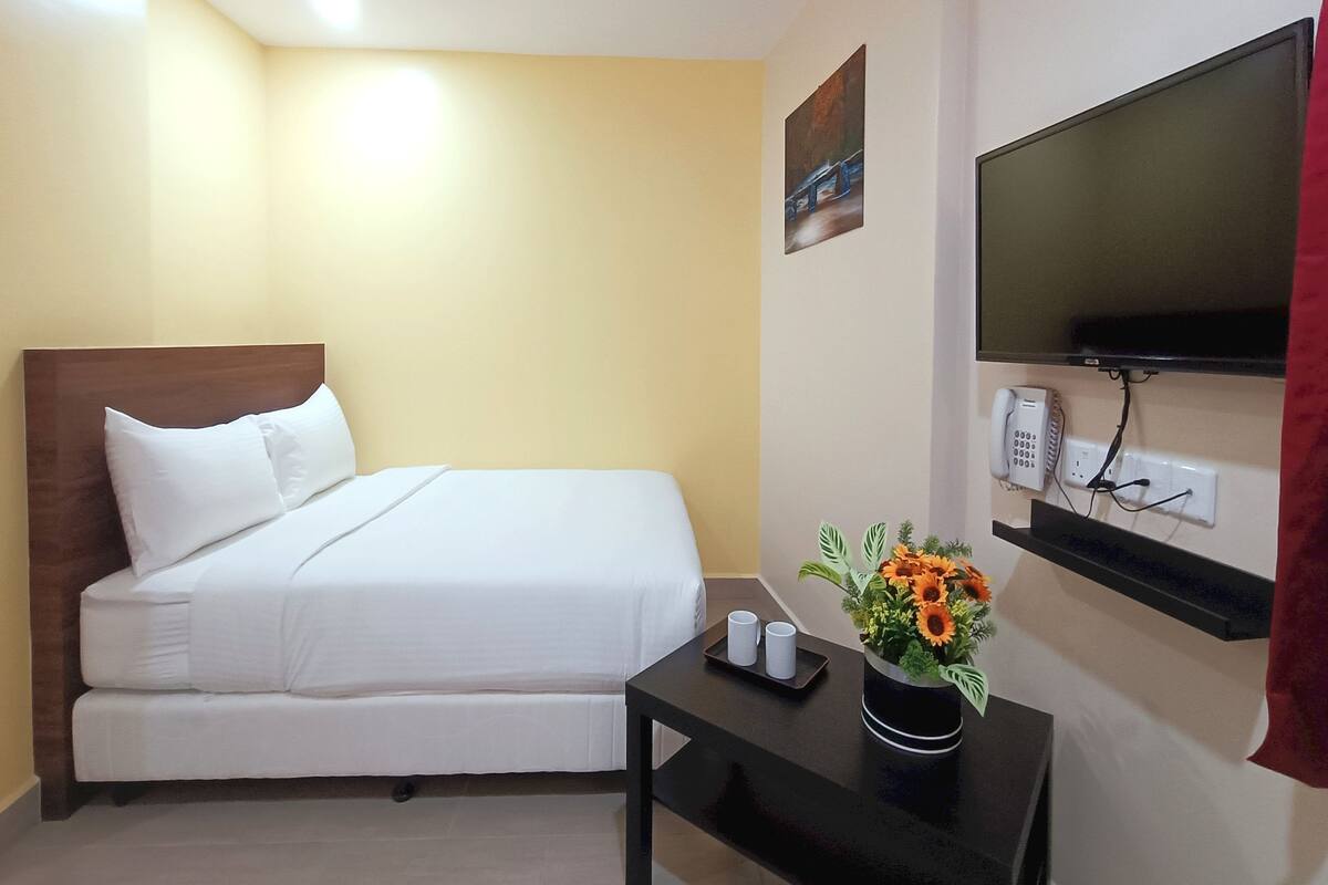 A hotel room with a bed, a small table with a vase of flowers, a TV mounted on the wall, and a telephone.