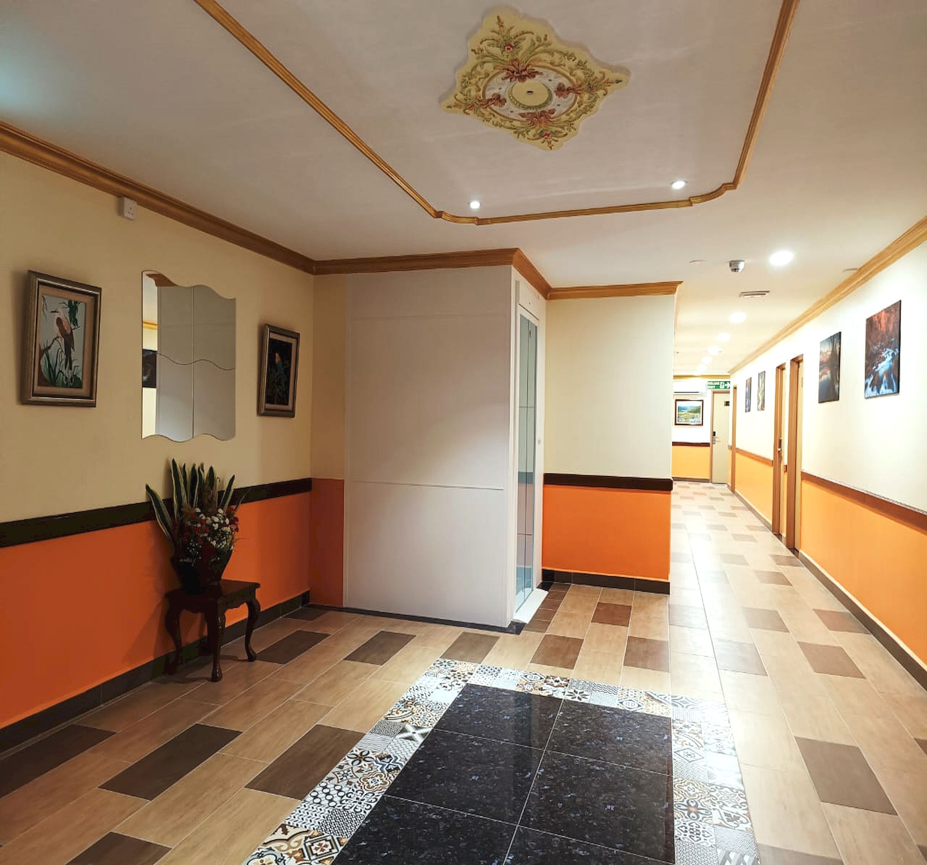 A hotel corridor with a patterned floor, a potted plant, framed artwork, and a decorative ceiling.