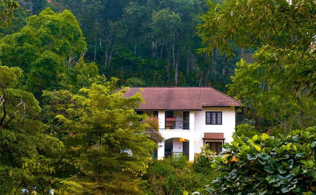 Una casa de dos pisos con techo de tejas rojas, rodeada de abundante follaje verde.