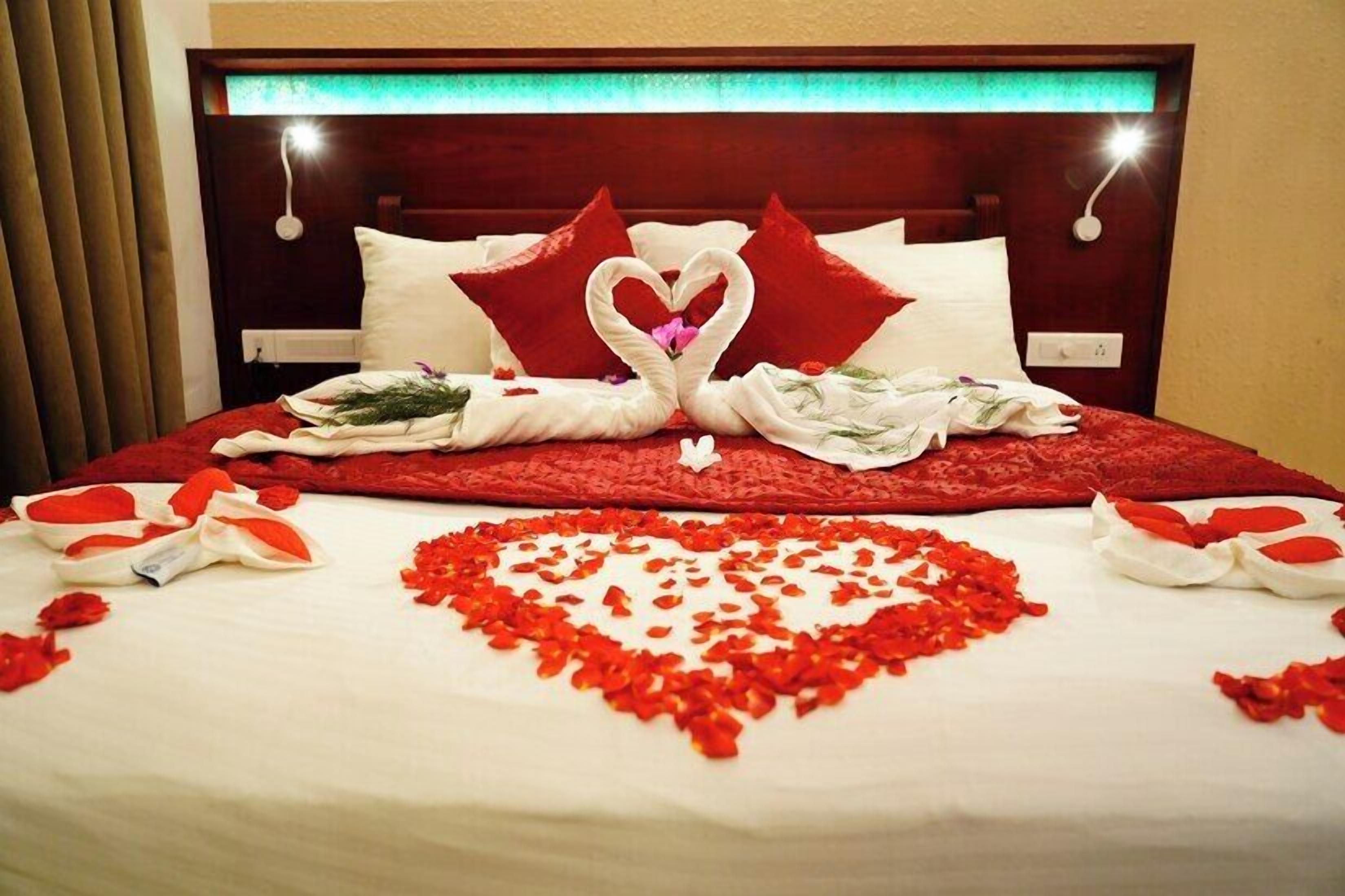 A bed with a heart-shaped arrangement of rose petals, a swan-shaped towel, and red pillows.