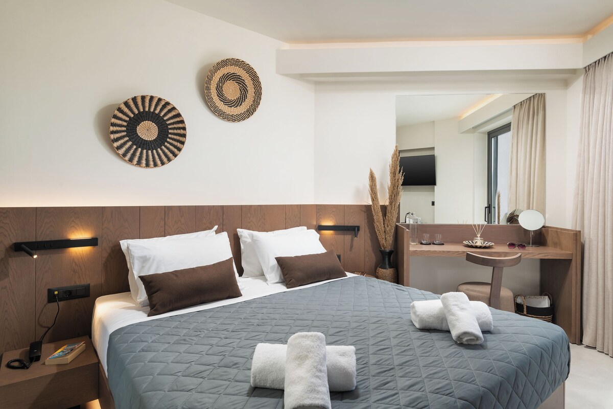 A modern hotel room with a large bed, a bedside table with a book, a wall-mounted mirror, and a small desk with a lamp.