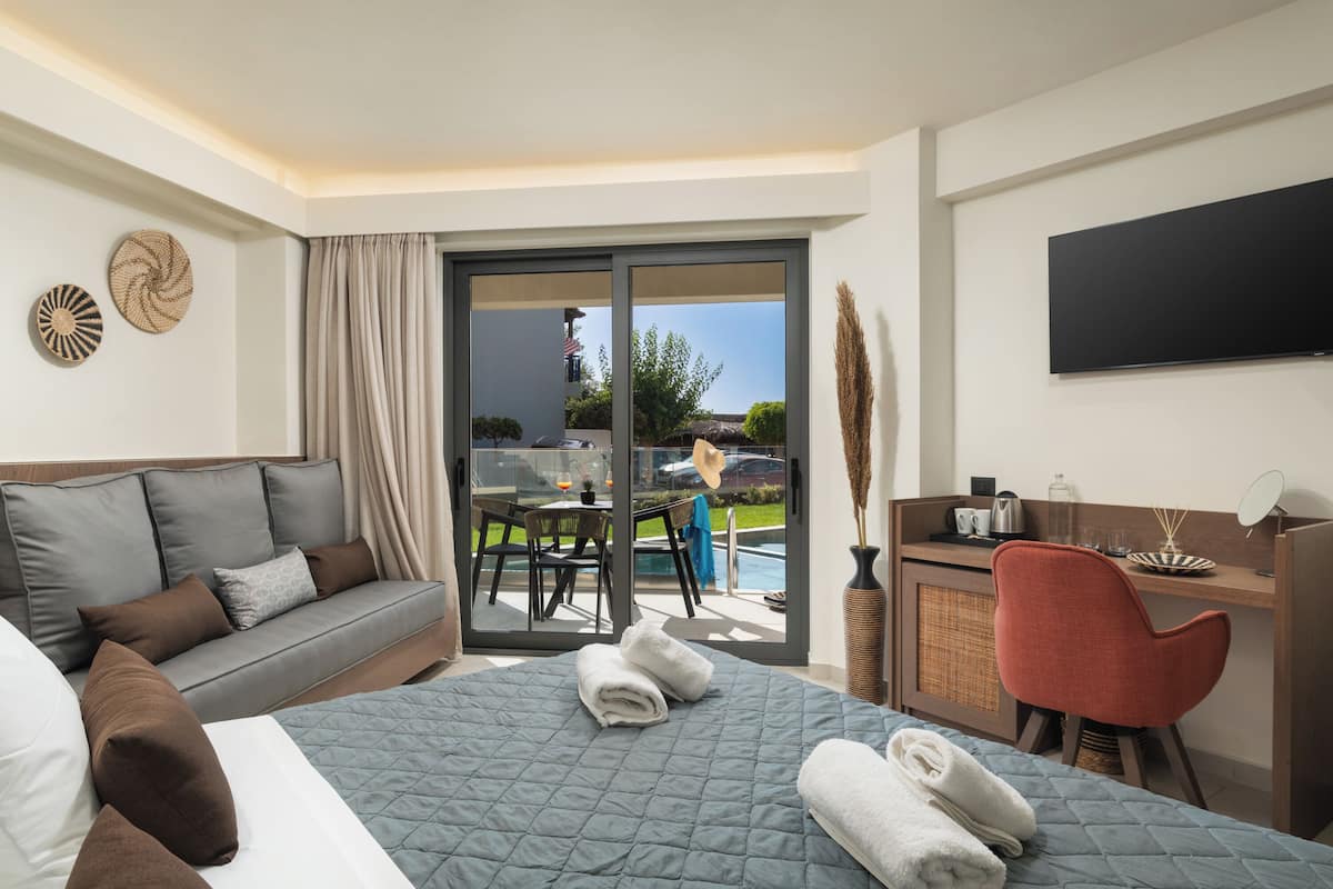 Une chambre d’hôtel comprenant un lit, un canapé, un bureau, une chaise, une télévision et une porte-fenêtre coulissante donnant sur un espace extérieur avec une piscine et une table.
