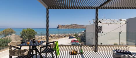 A balcony with a view of the sea, a table with chairs, and a basket of fruit.