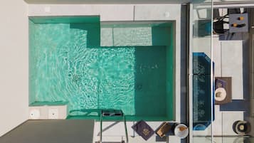Una piscina en la azotea con barandilla de cristal, una tumbona y un pequeño rincón de estar con un frutero y un libro.