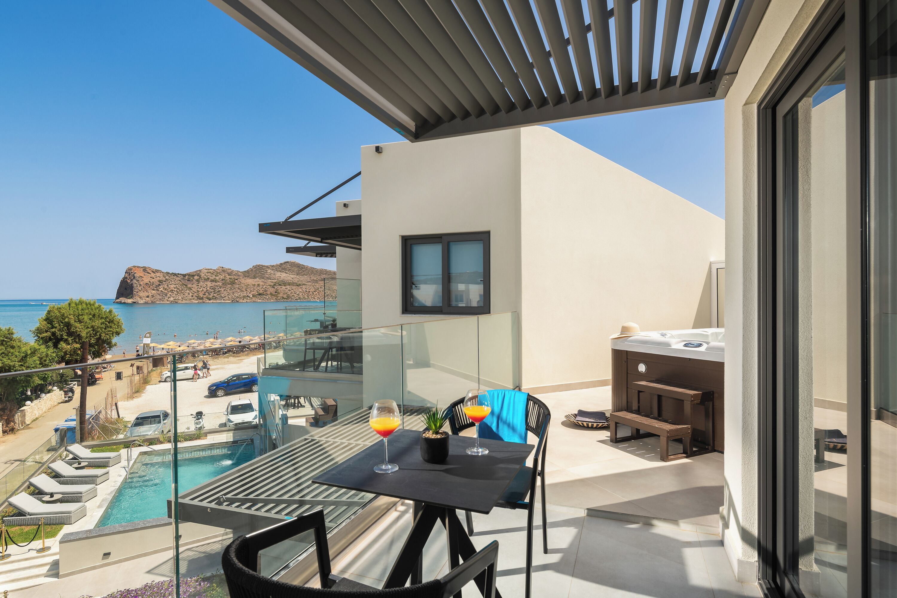 Un balcón con piscina, dos sillas, una mesa con bebidas y vistas al mar y a las montañas.