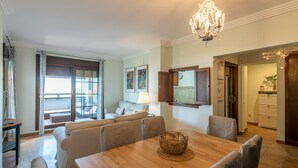 A dining area with a wooden table, chairs, and a chandelier. There is a sofa and a window with curtains in the background. - Las Palmeras - Punta del Moral Beach PLHP28P11 (Punta del Moral)