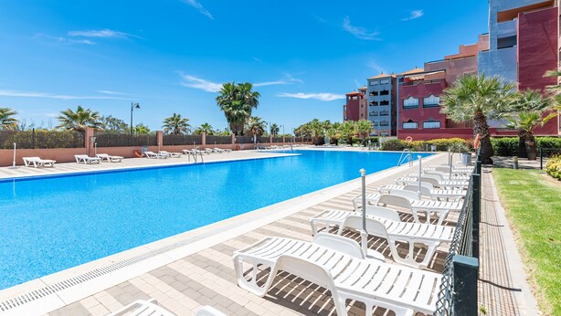 A swimming pool with lounge chairs, palm trees, and a red building in the background. - Las Palmeras - Punta del Moral Beach PLHP28P11 (Punta del Moral)