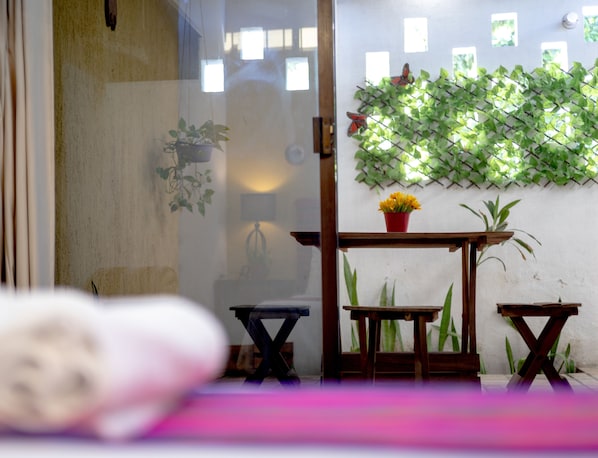 A room with a glass partition, a small wooden table with a red pot of flowers, and a green leafy wall decoration.