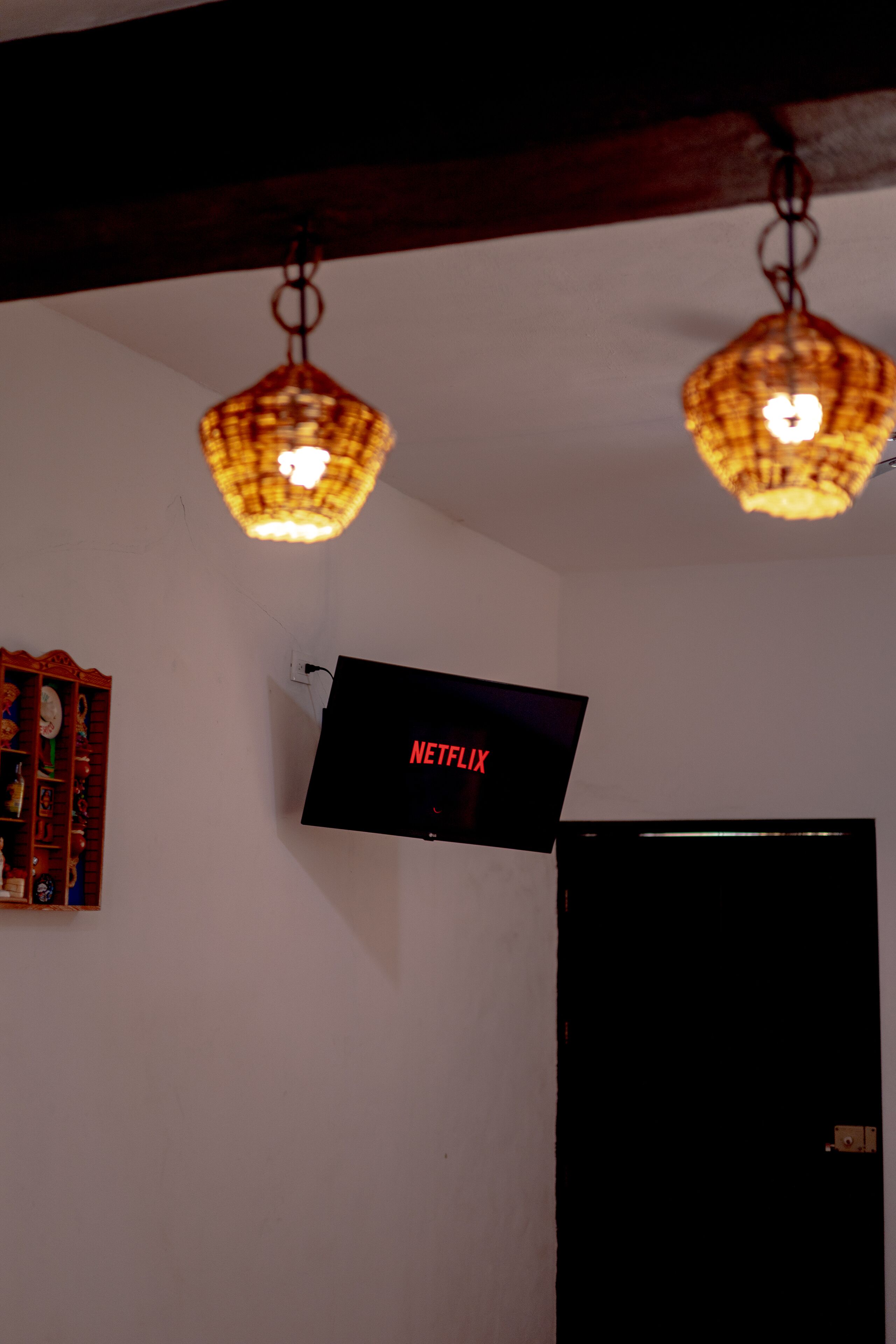 Una habitación con un televisor de pared que muestra 'NETFLIX' en letras rojas, una estantería de madera con varios objetos y dos lámparas colgantes.