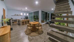 A modern interior with a staircase, a dining area with a round table and chairs, and a kitchen area with a countertop and hanging lights. - Villa Seseh Breeze (Cemagi)