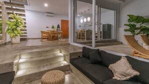 A modern living room with a dark sofa, a woven ottoman, and a staircase leading to a dining area with wooden furniture. - Villa Seseh Breeze (Cemagi)