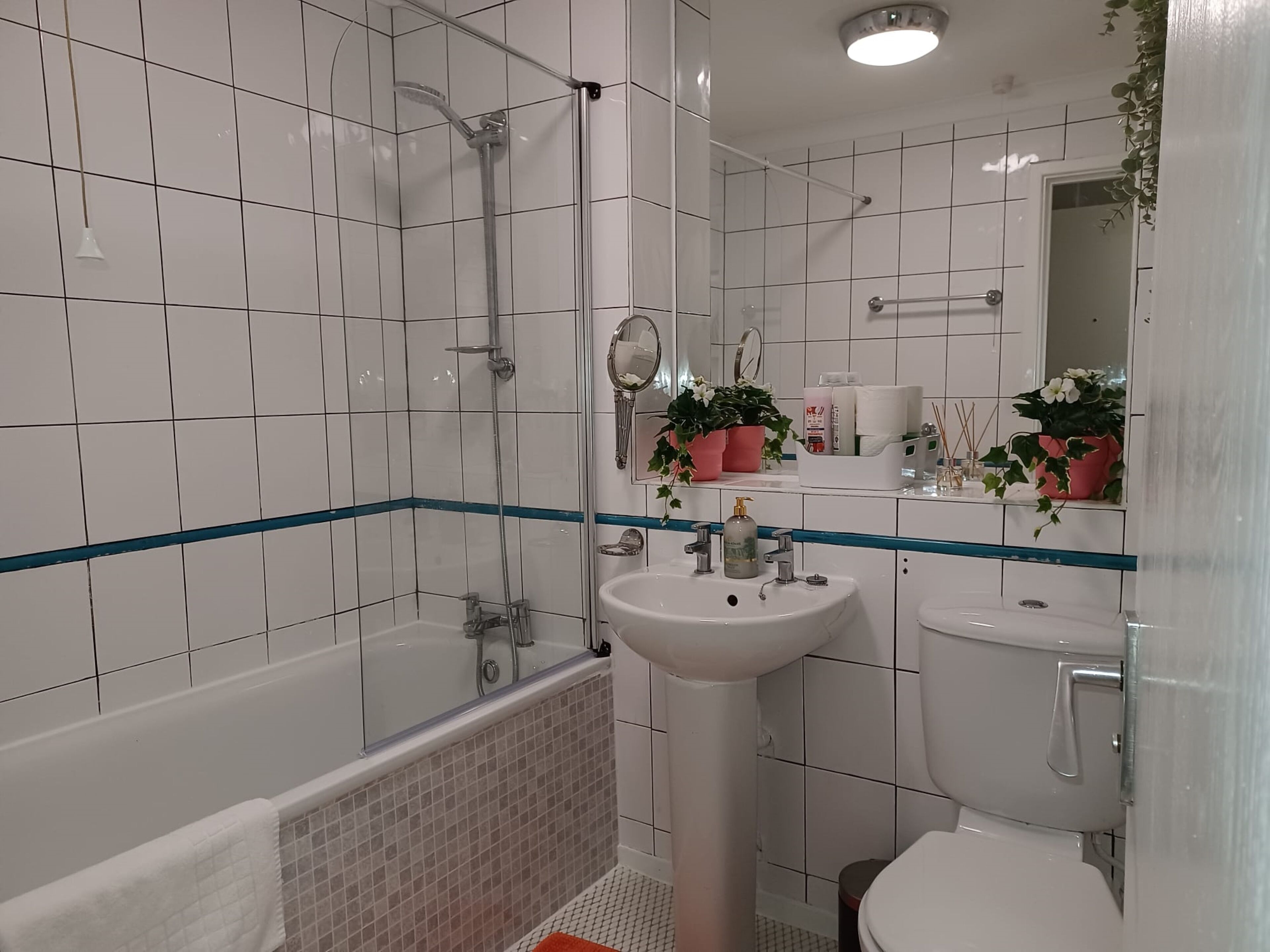 A bathroom with white tiled walls, a glass-enclosed shower, a white sink, and a toilet. There are potted plants on the vanity and a towel rack with a white towel.