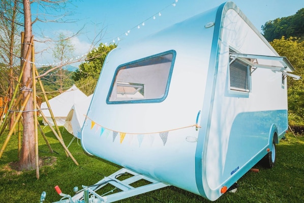 A vintage-style RV with a large window and a small attached structure, parked on grass with string lights. - kidsvillage (Dahu)