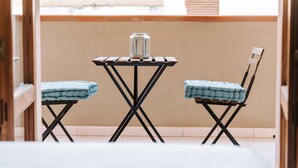 Un balcón con una mesa y dos sillas, un recipiente metálico sobre la mesa, y una vista de un edificio al fondo.