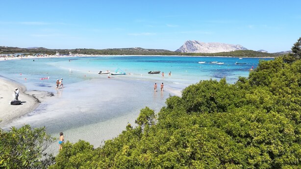 Una playa con aguas turquesas cristalinas, embarcaciones y montañas al fondo.