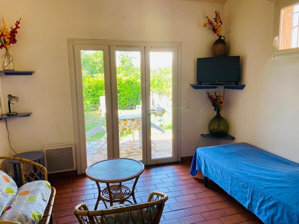 A room with a bed, a table, a chair, a TV, and a door leading to a garden.