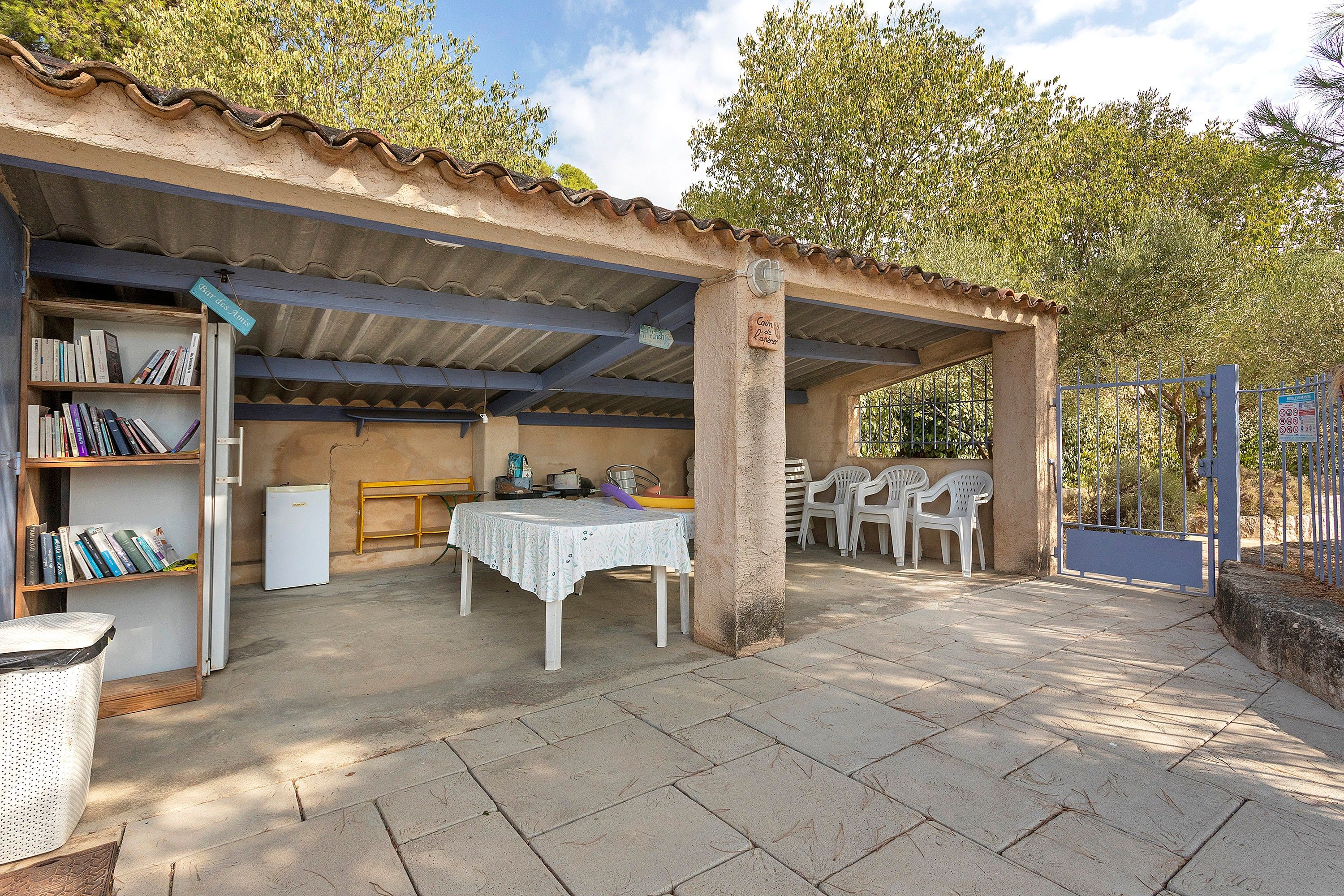 A covered outdoor area with a table and chairs, a bookshelf, and a refrigerator.