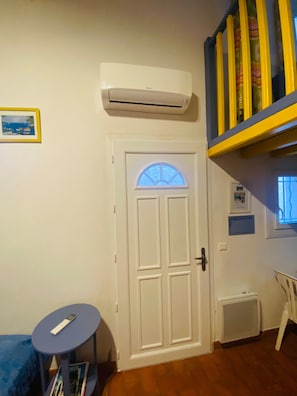 A room with a white door, an air conditioning unit above it, a small round table with a remote control, and a bookshelf with books.