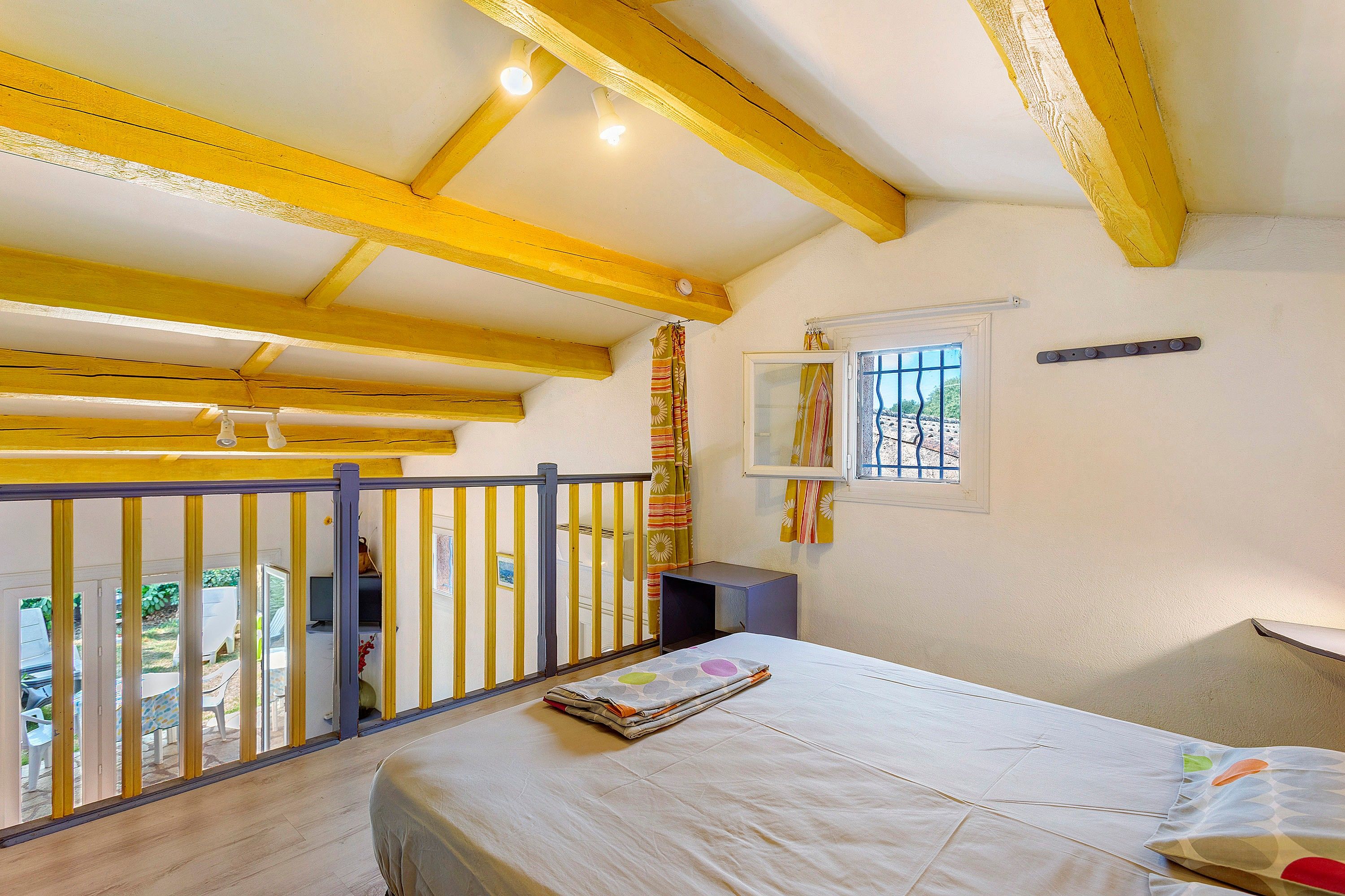 A room with a bed, a window with bars, a small table, and a balcony with yellow railings.