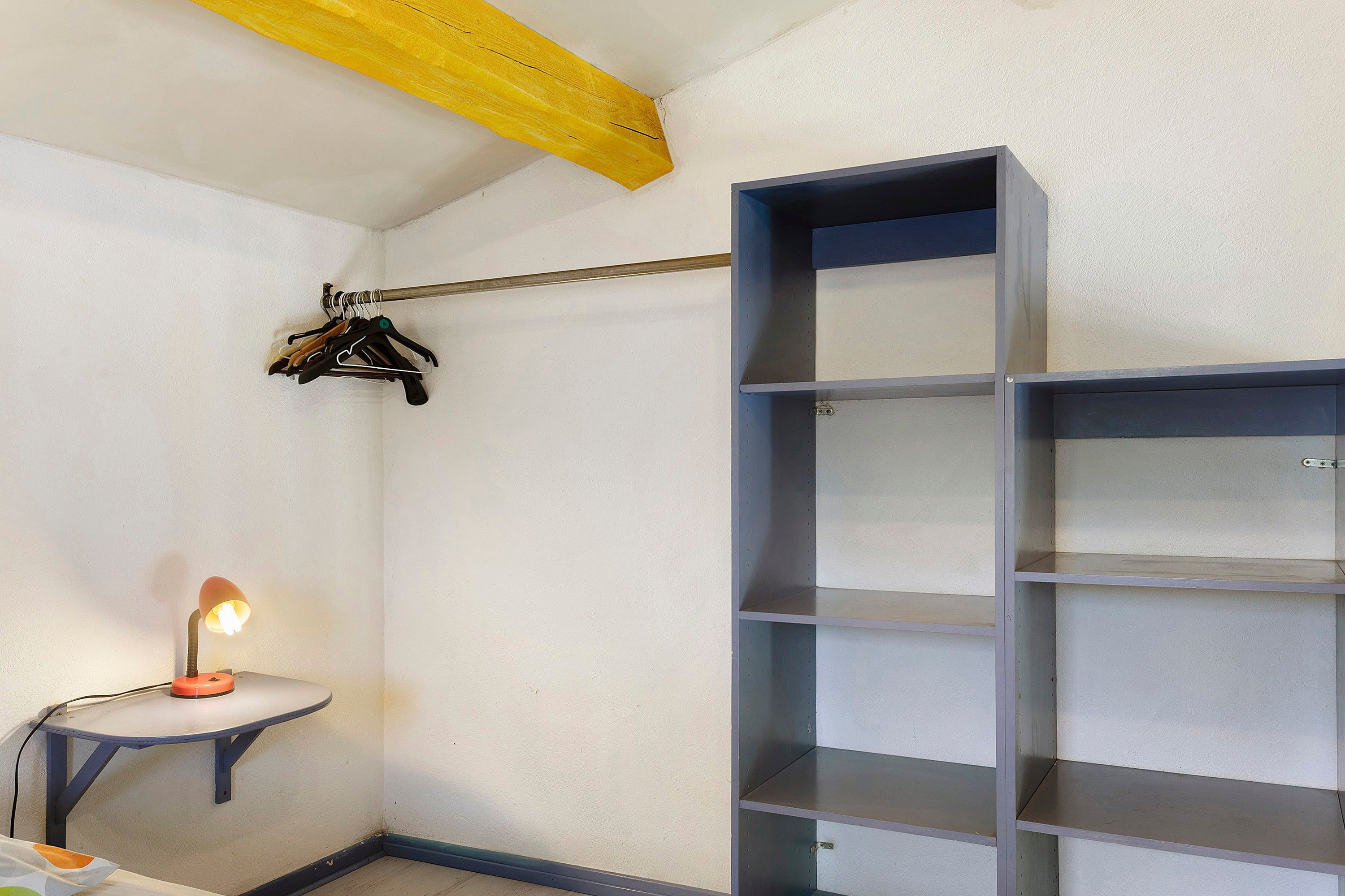 Un coin de pièce avec une étagère en métal, une petite table avec une lampe et un porte-manteau avec des vêtements.