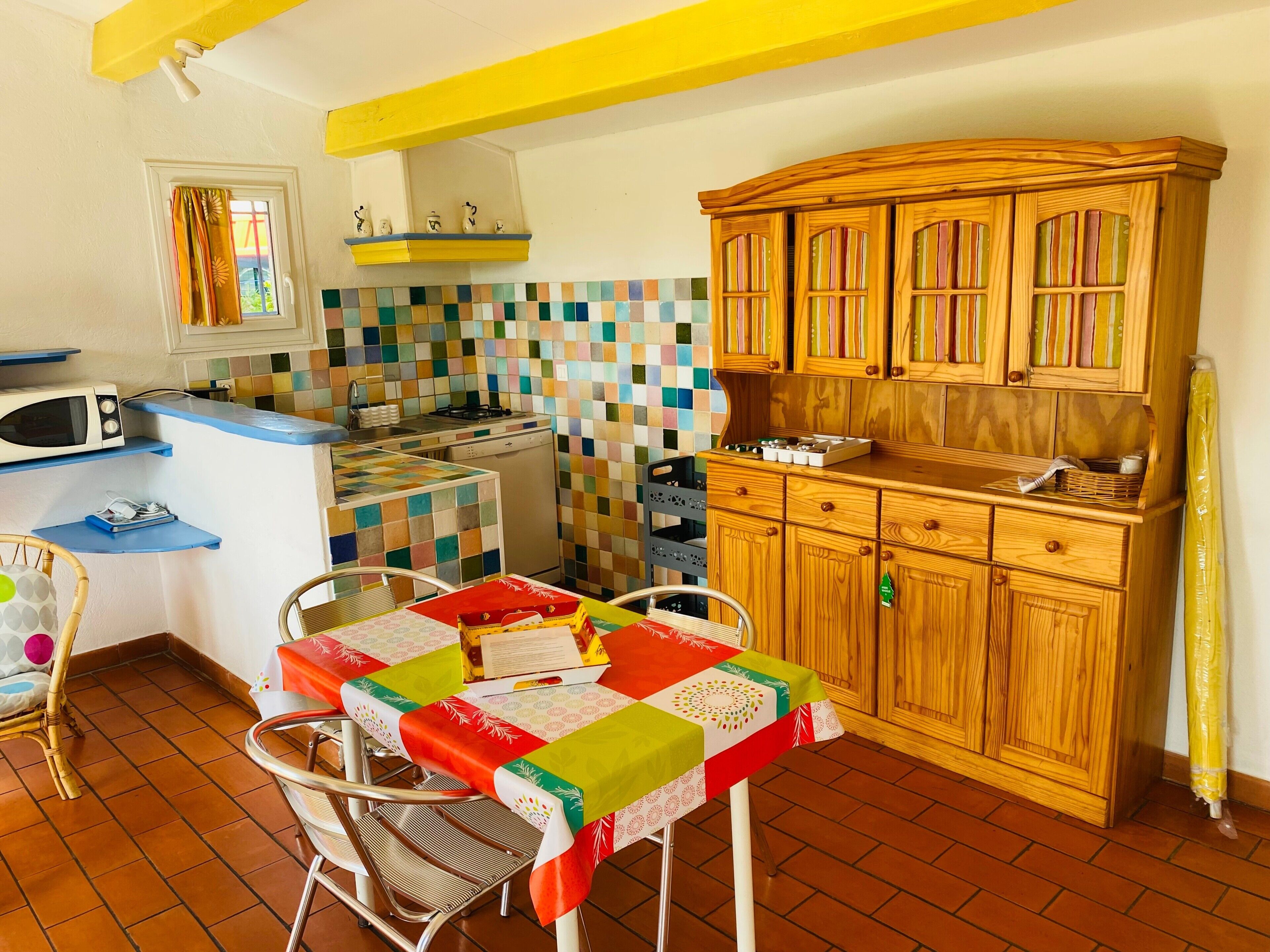 Une cuisine avec un mur recouvert de carreaux, des meubles en bois, un four à micro-ondes et une table recouverte d’une nappe colorée.
