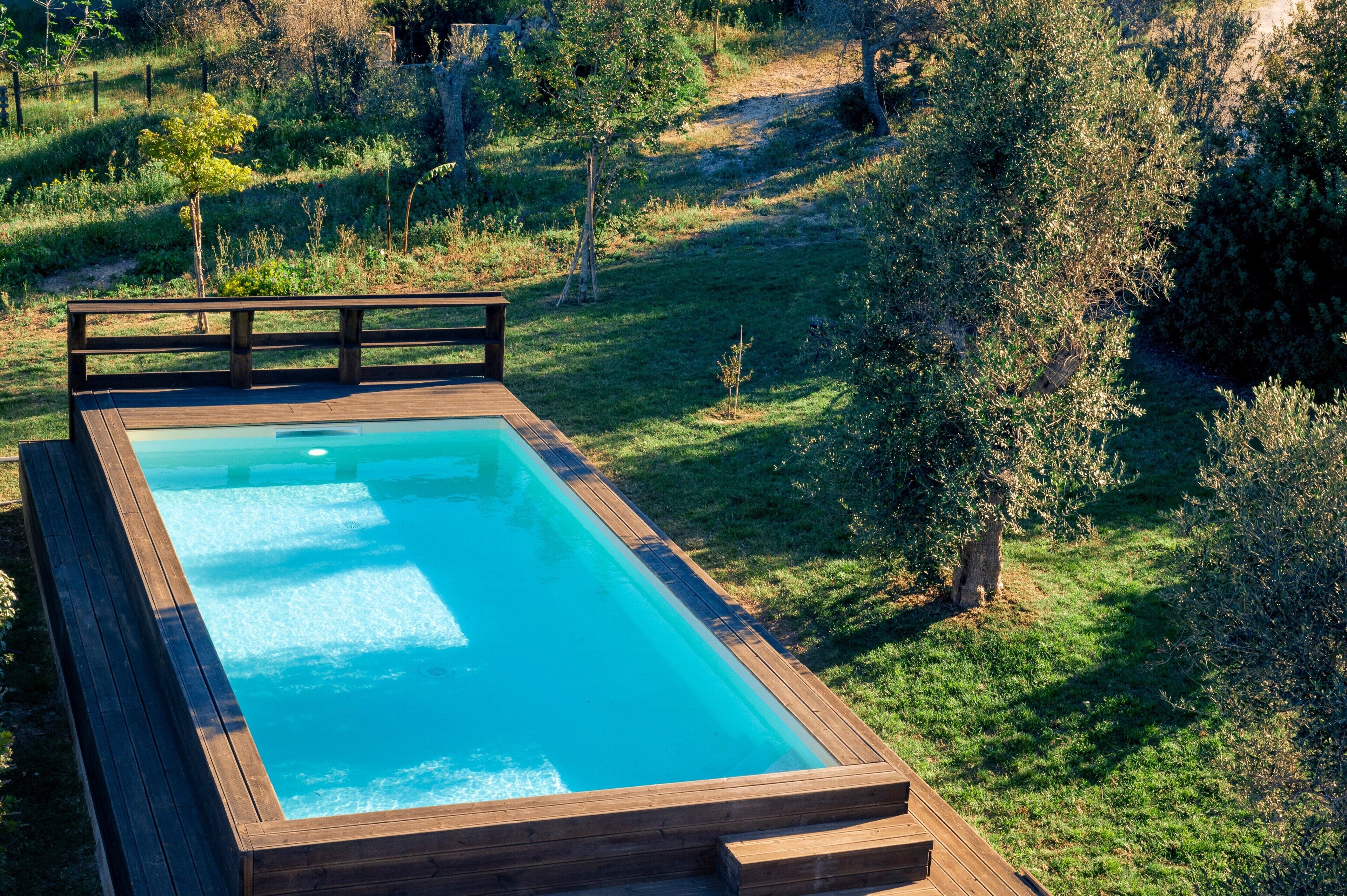 A rectangular pool with a wooden deck and a wooden railing, surrounded by a grassy area with trees and shrubs.