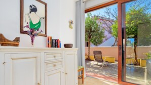 A room with a white cabinet, a framed artwork, a small table with books, and a view of an outdoor patio with a chair and a tree.