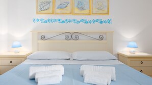 A neatly made bed with white linens and two folded towels, a wooden headboard with a decorative design, bedside lamps, and a wall with framed artwork and a floral border.