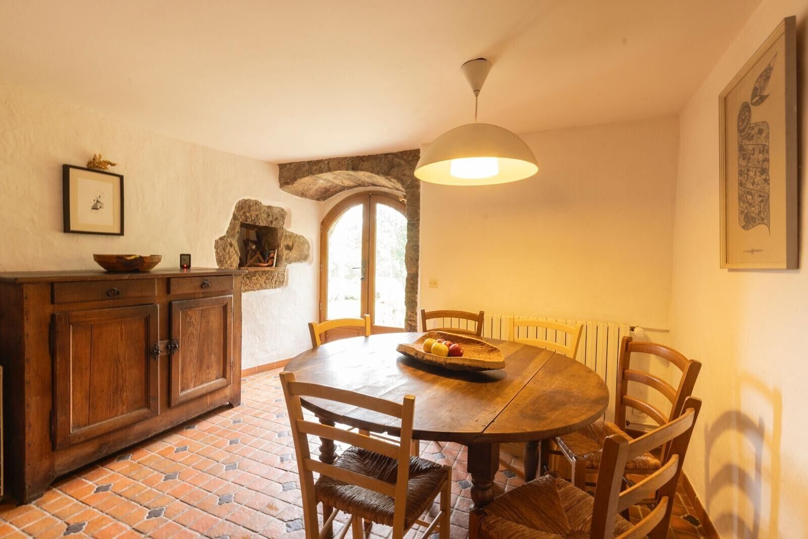 Un comedor con una mesa redonda de madera, seis sillas, una lámpara colgante, una chimenea de piedra y piso de baldosas.