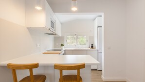 A modern kitchen with white cabinetry, a central island with two wooden chairs, and a view into the dining area. - Holiday Home "Casita Palmeras" with Mountain Views, Private Garden and Wi-Fi (Canyelles)