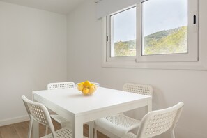 A white dining table with four chairs, a bowl of lemons, and a window with a view of a hill. - Holiday Home "Casita Palmeras" with Mountain View, Private Garden & Wi-Fi (Canyelles)