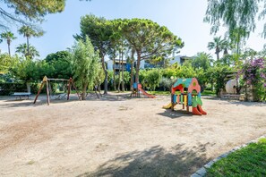 Un'area giochi con altalene e scivoli, circondata da alberi e edifici.