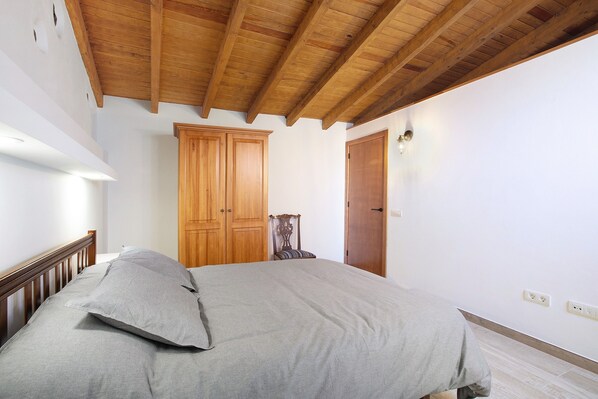 Un dormitorio con techo de madera, cama con ropa de cama gris, armarios de madera y una silla.