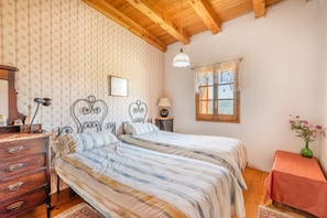 A bedroom with two beds, wooden ceiling beams, a window with lace curtains, a dresser, and a vase with flowers.