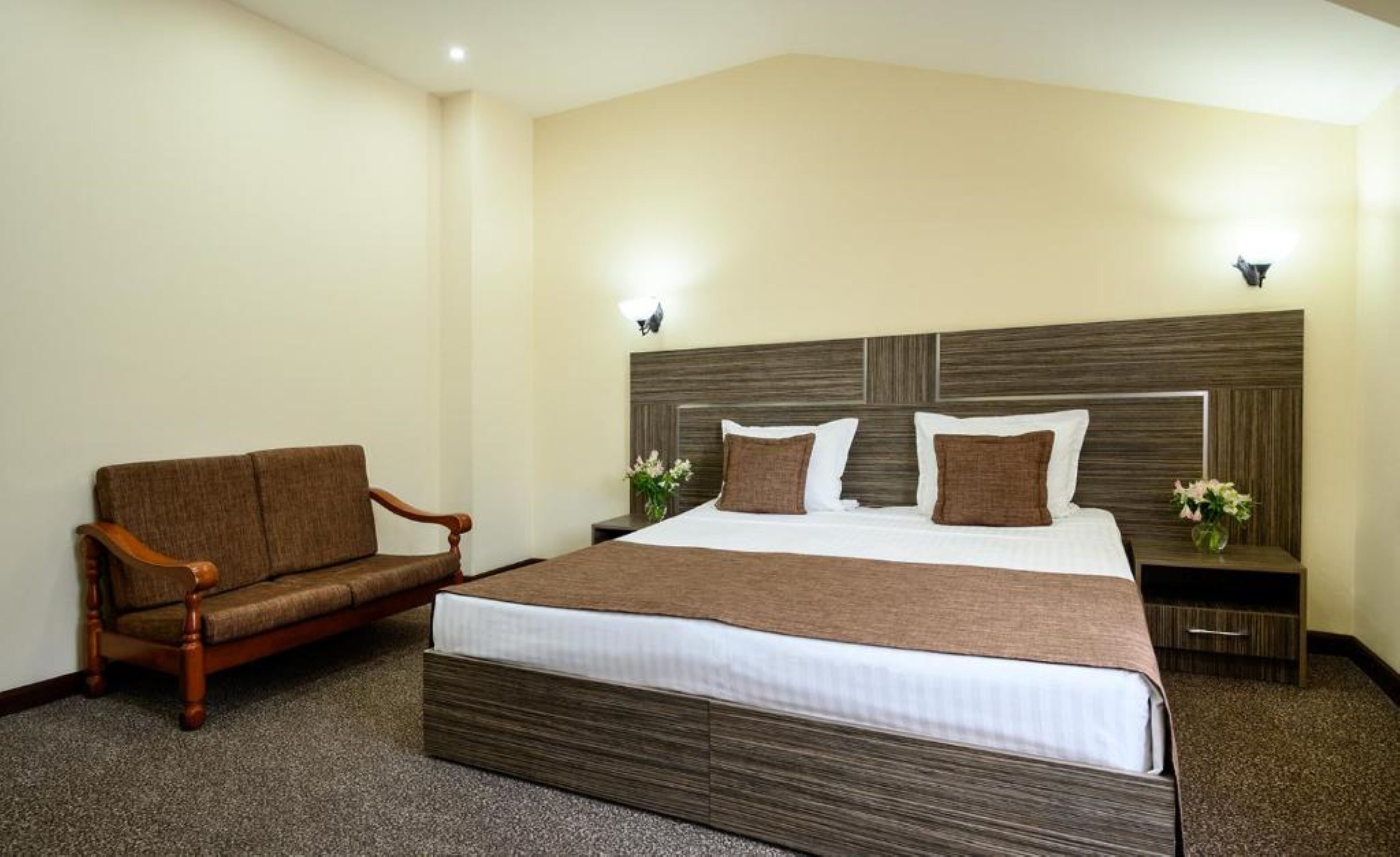 Een hotelkamer met een groot bed, een houten fauteuil, twee nachtkastjes met lampen, en een bijzettafeltje met een bloevas.