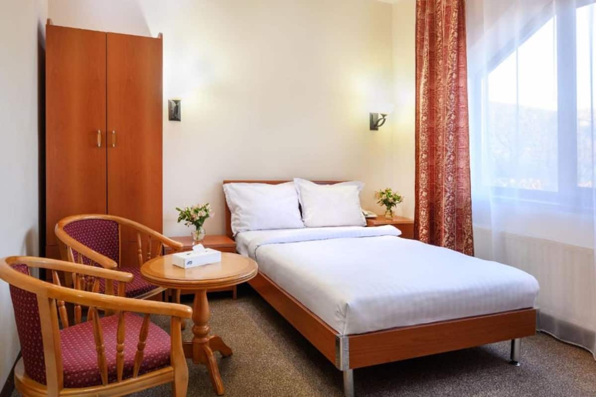 Una habitació d'hotel amb un llit, un armari de fusta, una taula rodona amb una cadira i una finestra amb cortines.